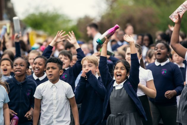 A diverse crowd of happy schoolchildren in England waving reusable water bottles and running towards the camera.
