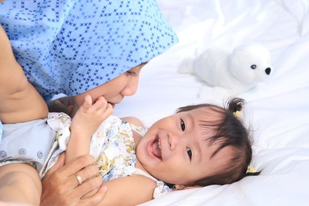 A happy, laughing baby wearing a reusable diaper or nappy and lying on a bed which its mother, in a headscarf, plays with it.
