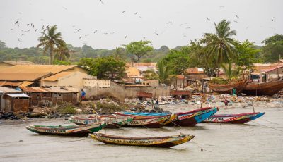 An aerial photo of the Gambian coast looking towards boats and palm trees. The beach is covered with brightly-coloured plastic pollution.
