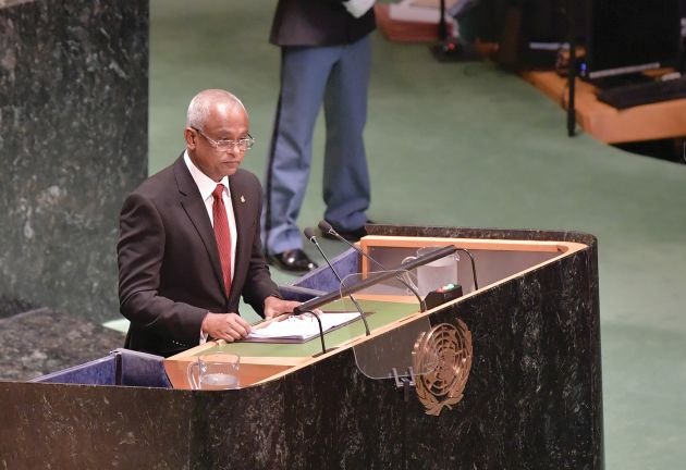 A Maldivian man, President Ibrahim Mohamed Solih, speaks at the UN General Assembly. He is wearing a dark suit and glasses, standing behind a large black speaker's desk with the UN logo on the front.