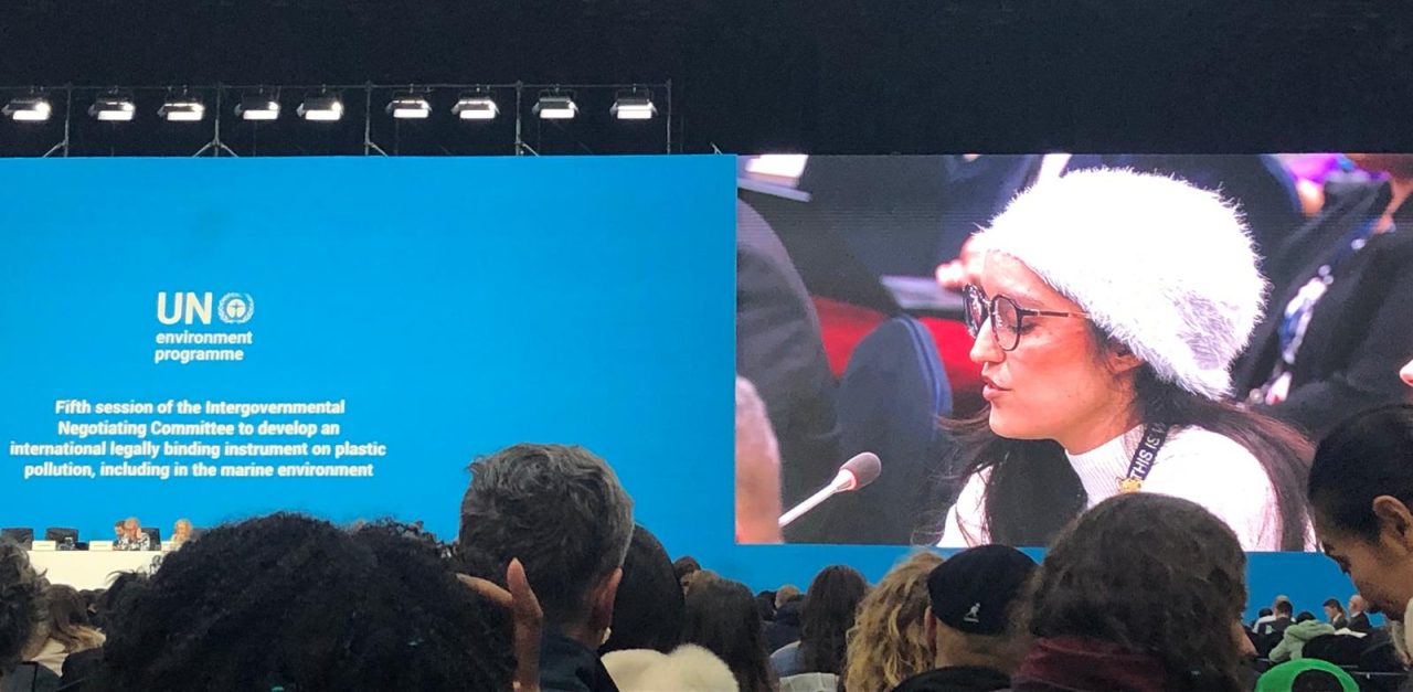 Delegate from Mexico makes a speech at the closing plenary of INC5 in Busan. In the foreground is the crowd, and on the screen is the blue INC sign and a shot of the delegate, wearing a light top and a light fluffy bucket hat.