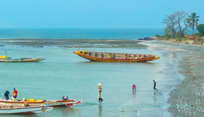 The coast of The Gambia, showing fishing boats in the shallow coastal seas, and a man carrying a box from teh boat to the shore. On the horizon, we can see the mid-blue Atlantic, with trees on the right side of the photo.