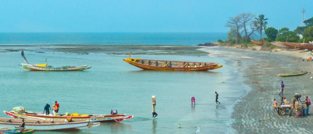 The coast of The Gambia, showing fishing boats in the shallow coastal seas, and a man carrying a box from teh boat to the shore. On the horizon, we can see the mid-blue Atlantic, with trees on the right side of the photo.