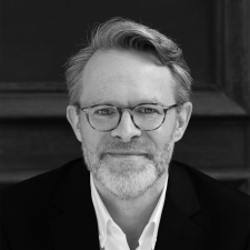 Profile photo of James Simpson, Head of Communications and Marketing at Common Seas. James wears glasses, a white shirt and dark blazer and smiles at the camera. Photo is in greyscale.